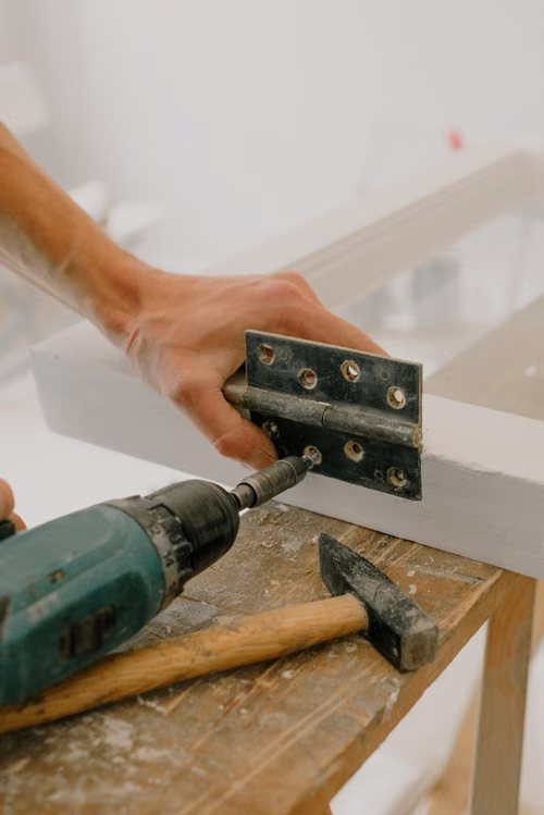 Drilling a hinge onto a painted wooden frame, with a hammer on the workbench